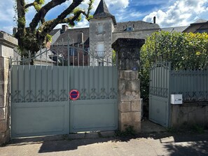 A gated entrance with a metal gate, a stone pillar, and a building with a tower in the background. - Manor house + swimming pool (Meyssac)