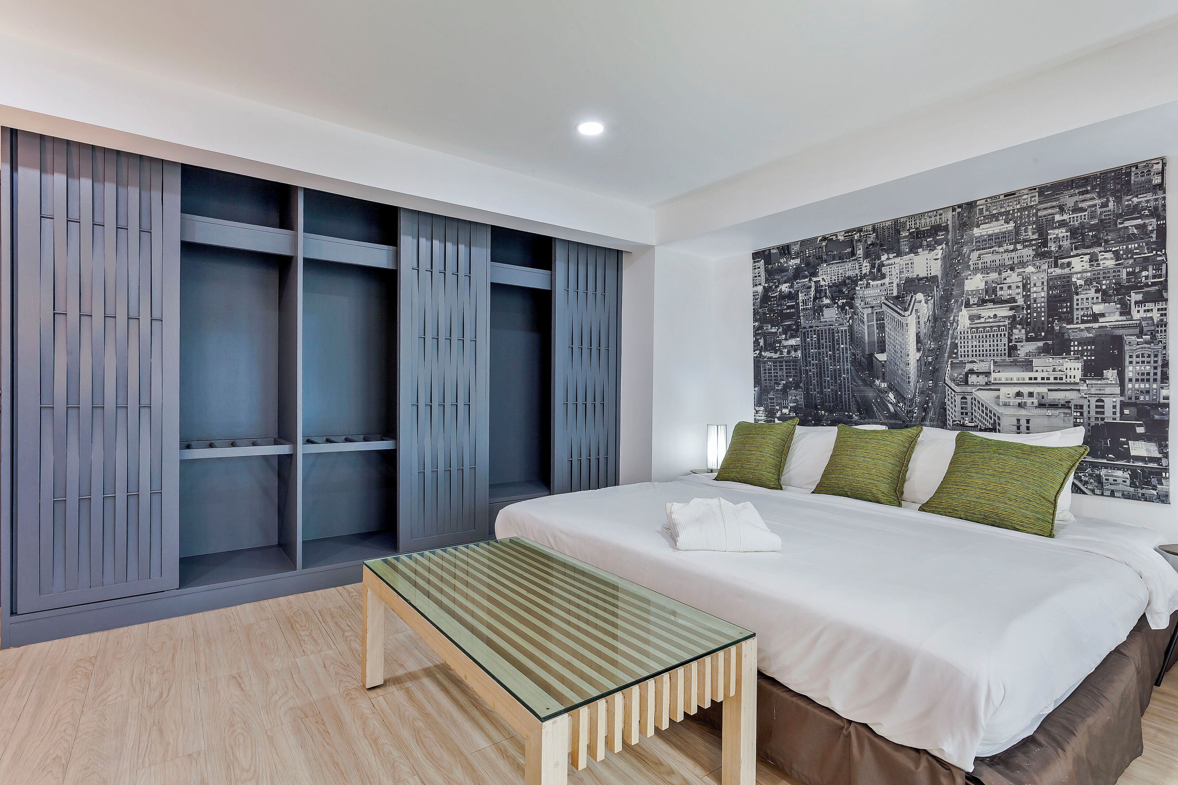 Ein Hotelzimmer mit einem Bett, einer Sitzbank, einem Kleiderschrank und einem Stadtblick an der Wand.