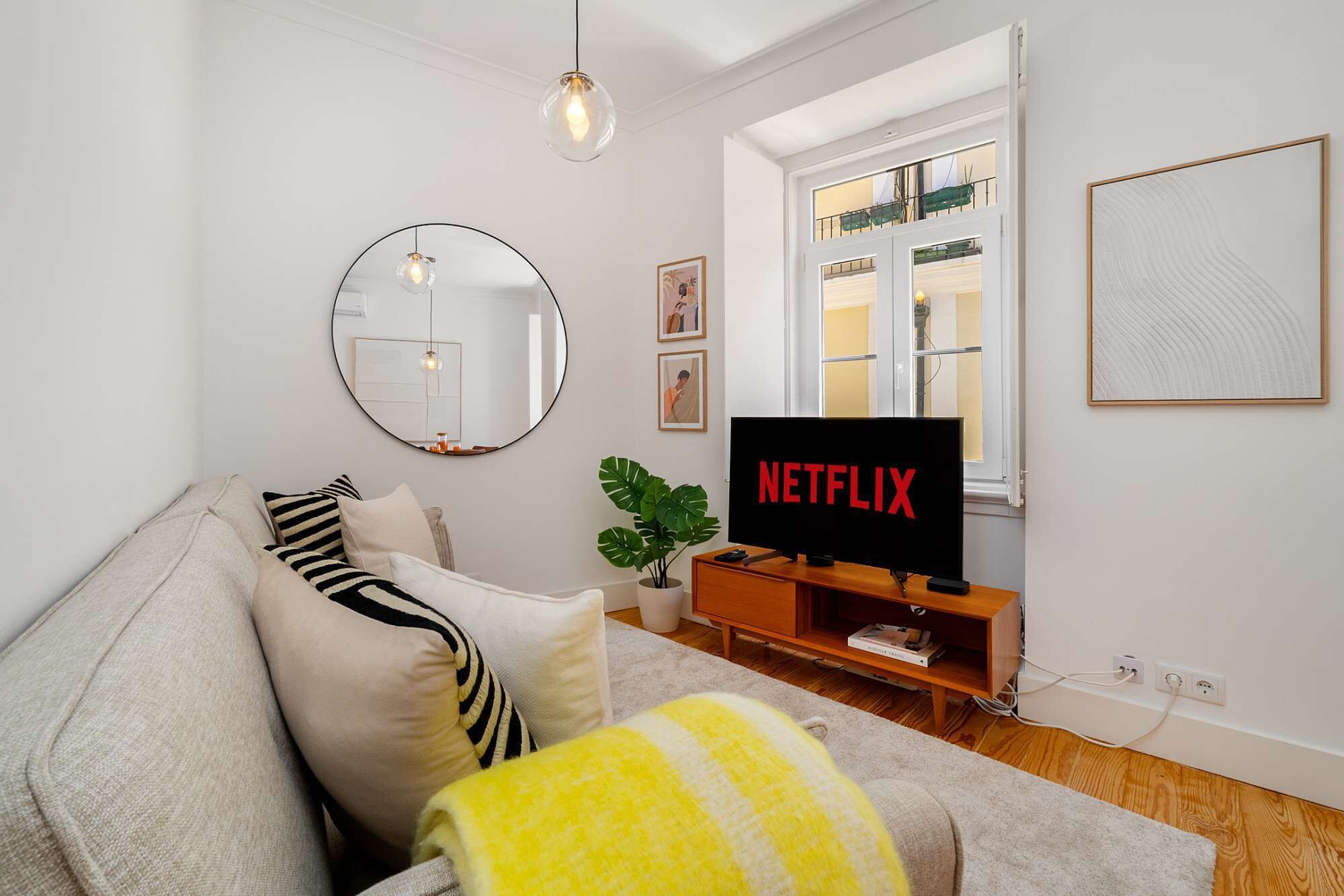A living room with a sofa, a round mirror, a television displaying 'NETFLIX,' and a window with blinds.