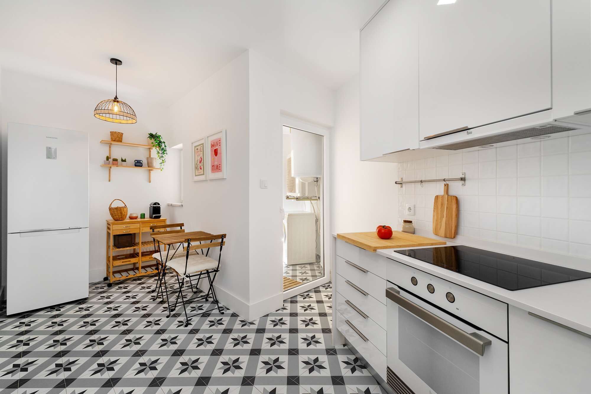 Una cocina moderna con muebles blancos, piso con estampado blanco y negro, una mesa de comedor de madera con sillas, y una lámpara colgante.