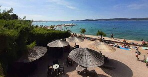 A beachside area with outdoor seating and umbrellas, a view of the sea, and a pier in the background. - Holiday apartment with garden and air conditioning (Turanj)