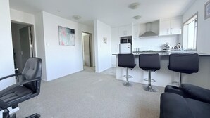 A modern kitchen with white cabinets, a microwave, and a coffee machine. There are four black bar stools and a black office chair. The floor is carpeted, and the walls are white.