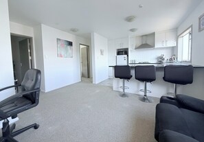 A modern kitchen with white cabinets, a microwave, and a coffee machine. There are four black bar stools and a black office chair. The floor is carpeted, and the walls are white.