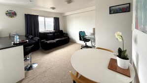 A modern living room with a black sofa, a dining table with chairs, a small kitchen area, and a window with curtains.