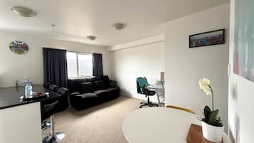 A modern living room with a black sofa, a dining table with chairs, a small kitchen area, and a window with curtains.