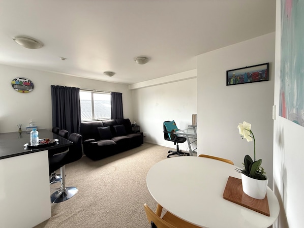 A modern living room with a black sofa, a dining table with chairs, a small kitchen area, and a window with curtains.