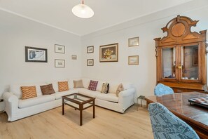 En stue med en hvid sofa, et træbord og et spisebord med stole. Væggen er dekoreret med billeder i rammer, og der står et træskab med glasdøre.
