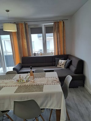 Ein modernes Wohnzimmer mit einer Couch, einem Esstisch und Stühlen. Vor den Fenstern hängen Vorhänge, und auf dem Tisch steht eine Flasche.