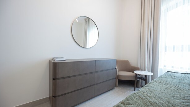 Una habitación de hotel moderna con un armario gris, un espejo redondo, una pequeña mesa redonda y una cama con una manta verde.