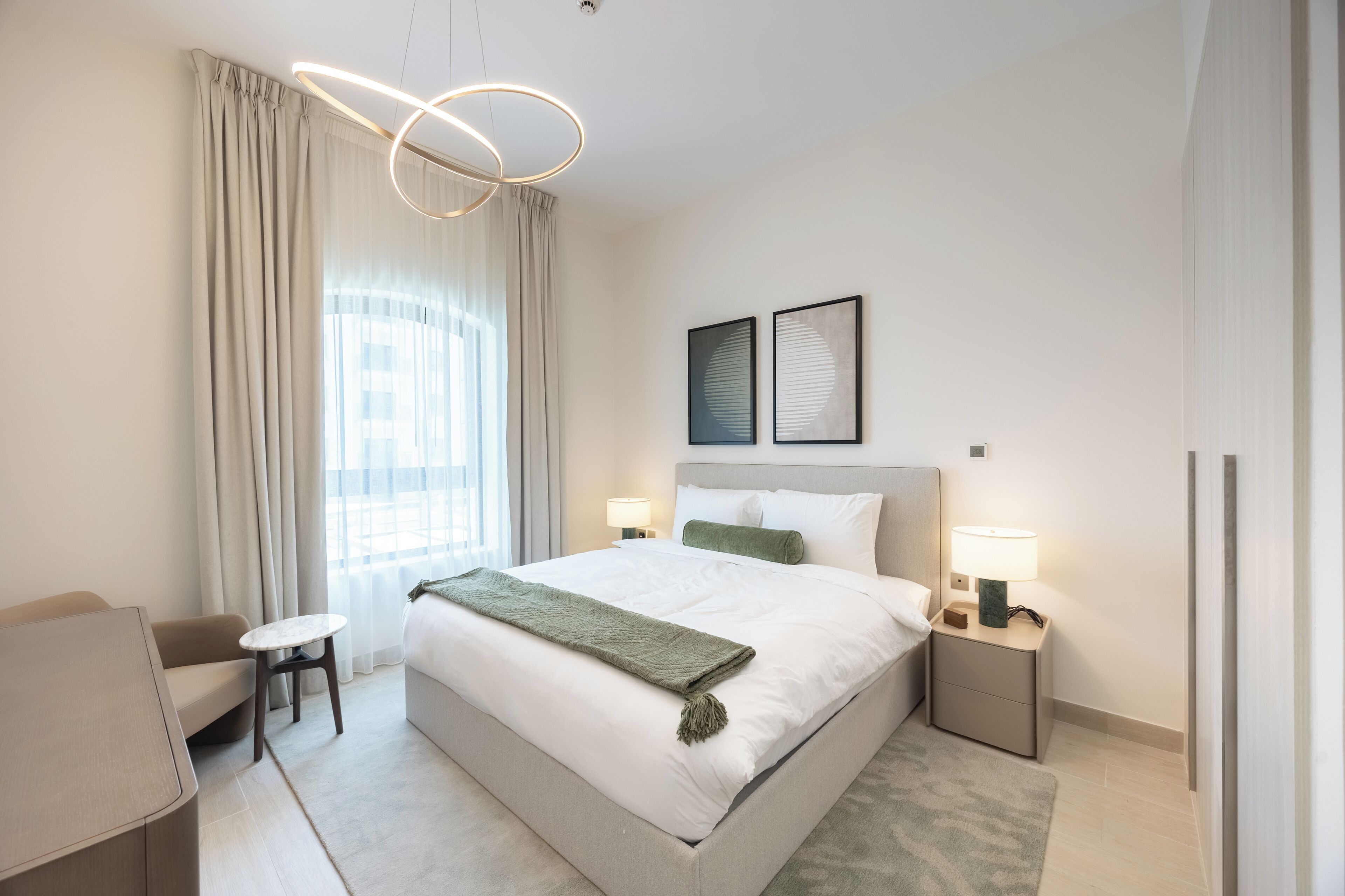 A modern hotel room with a large bed, bedside lamps, a small table, and a view of the city through the window.