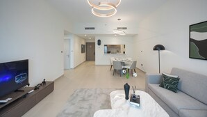 Una sala de estar moderna con una televisión de pantalla plana, una mesa de centro y un sofá. Se observa un comedor con una mesa y sillas al fondo.