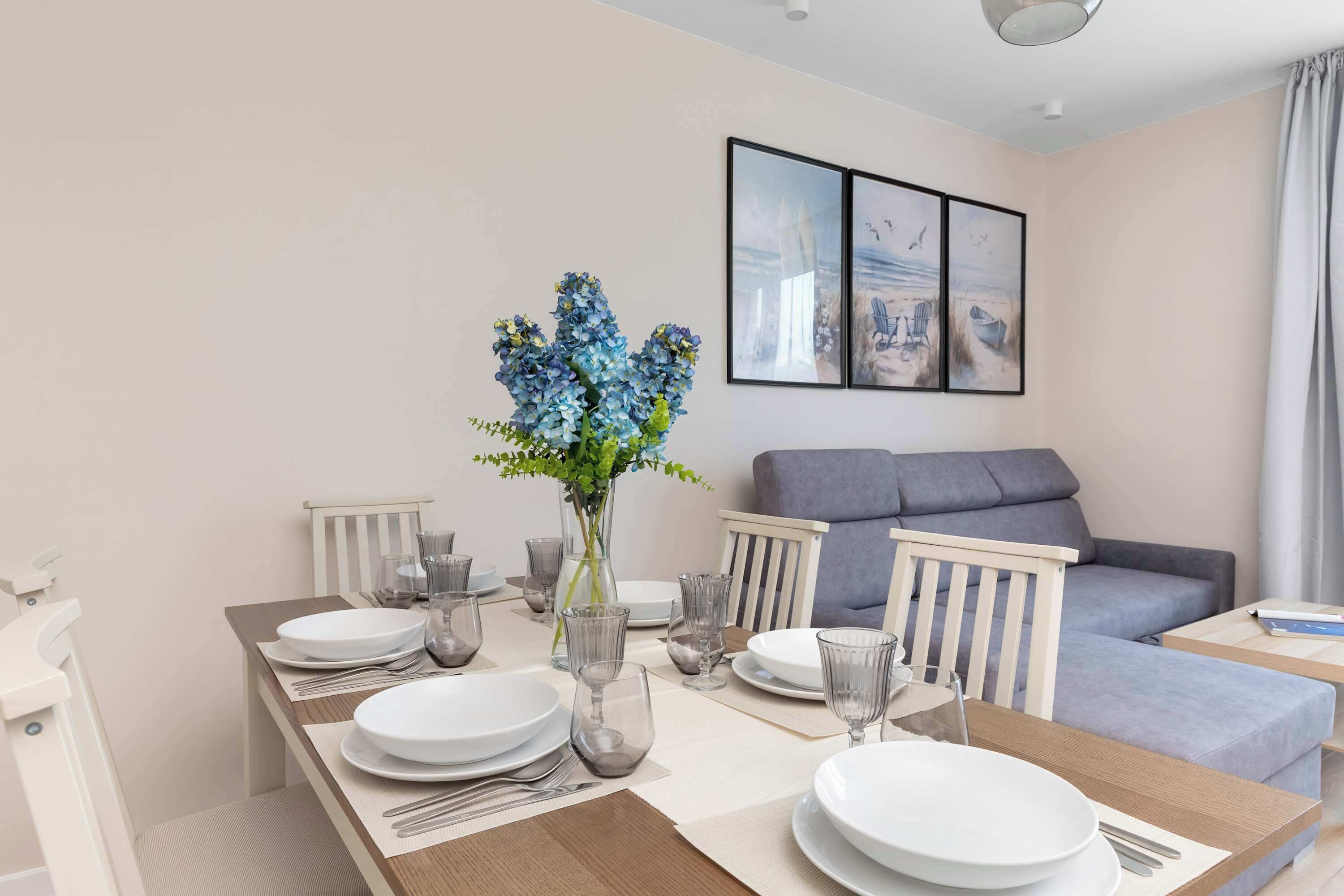 A dining area with a table set for four, a vase with blue flowers, and a grey sofa in the background.