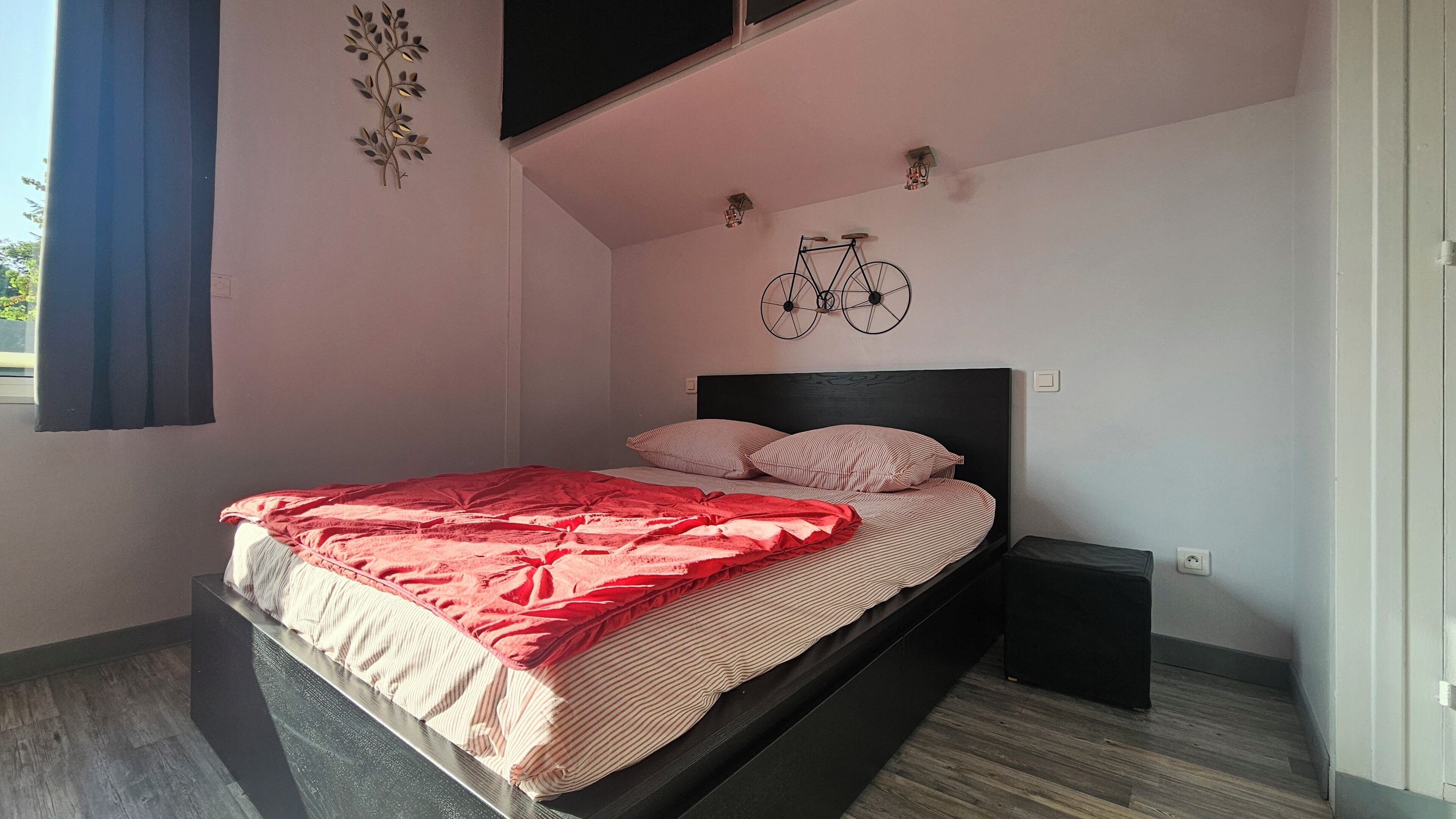 Een enkel bed met een rode deken en gestreepte kussens, een zwart hoofdbord en een fietsdecoratie aan de muur.