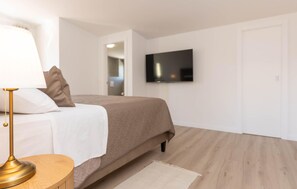 Un dormitorio con cama, lámpara de noche, televisión montada en la pared y piso de madera.