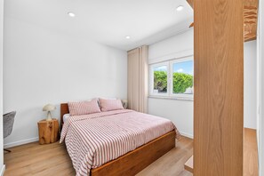 Ein Schlafzimmer mit einem hölzernen Bett, einem Nachttisch, einer Lampe und einem Fenster mit Blick auf Grünflächen.