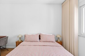 Ein Schlafzimmer mit einem Bett, zwei Nachttischen mit Lampen, einem Sessel und einem Fenster mit Vorhängen.