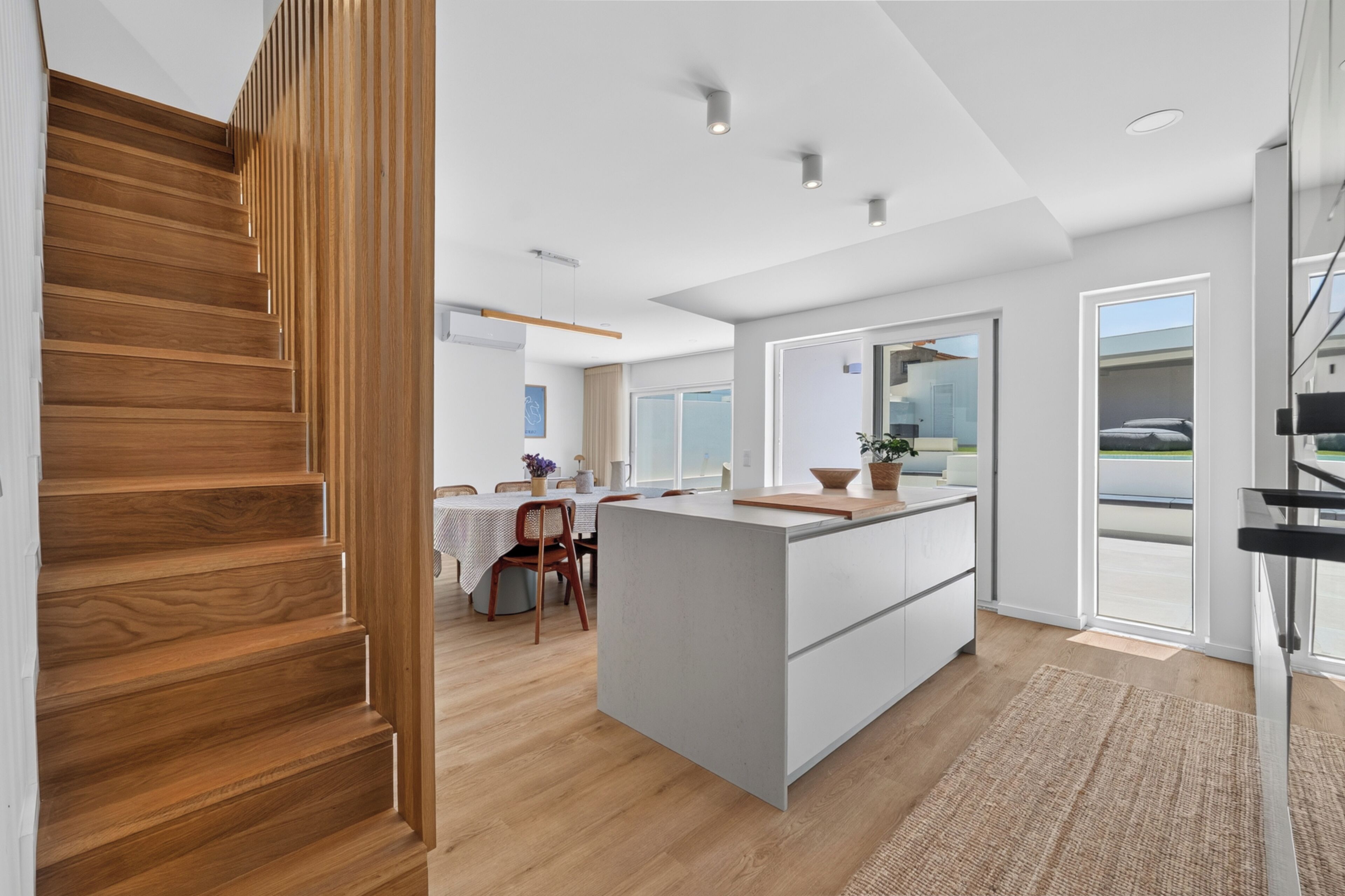 A modern kitchen with a central island, wooden staircase, and a dining area with a table and chairs.