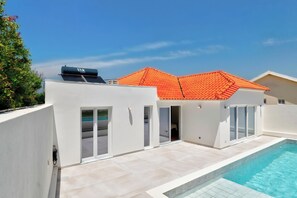 Ein moderner Poolbereich im Freien mit blauem Himmel, einem roten Ziegeldach und einem weißen Gebäude mit einer Glas-Schiebetür.