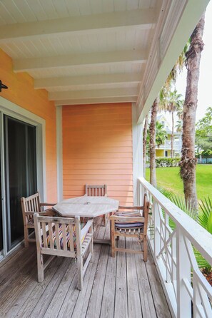 A wooden deck with a table and chairs, a white railing, and a view of palm trees and a building in the background.