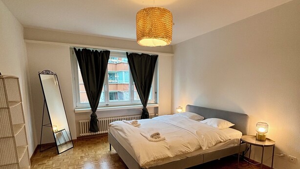 Un dormitorio con cama, espejo de pie, radiador, ventana con cortinas y lámpara colgante.