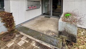 Entrada de un edificio con un área techada, panel de alarma contra incendios y una jardinera verde.