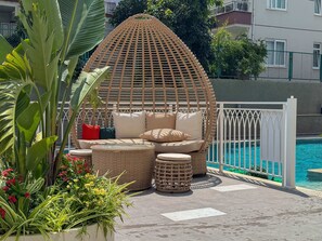 Una silla de mimbre con cojines, una mesita redonda y una piscina al fondo.