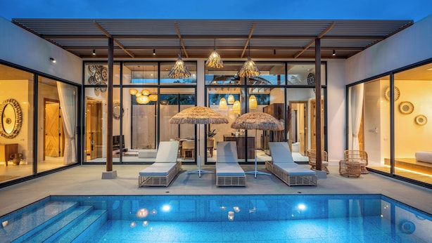 Uma área de piscina moderna ao ar livre, com espreguiçadeiras e guarda-sóis, cercada por paredes de vidro e uma pérgula de madeira.