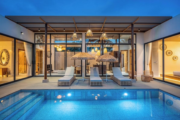 A modern outdoor pool area with lounge chairs and umbrellas, surrounded by glass walls and a wooden pergola. - Westkey Luxury Villa (Kathu)
