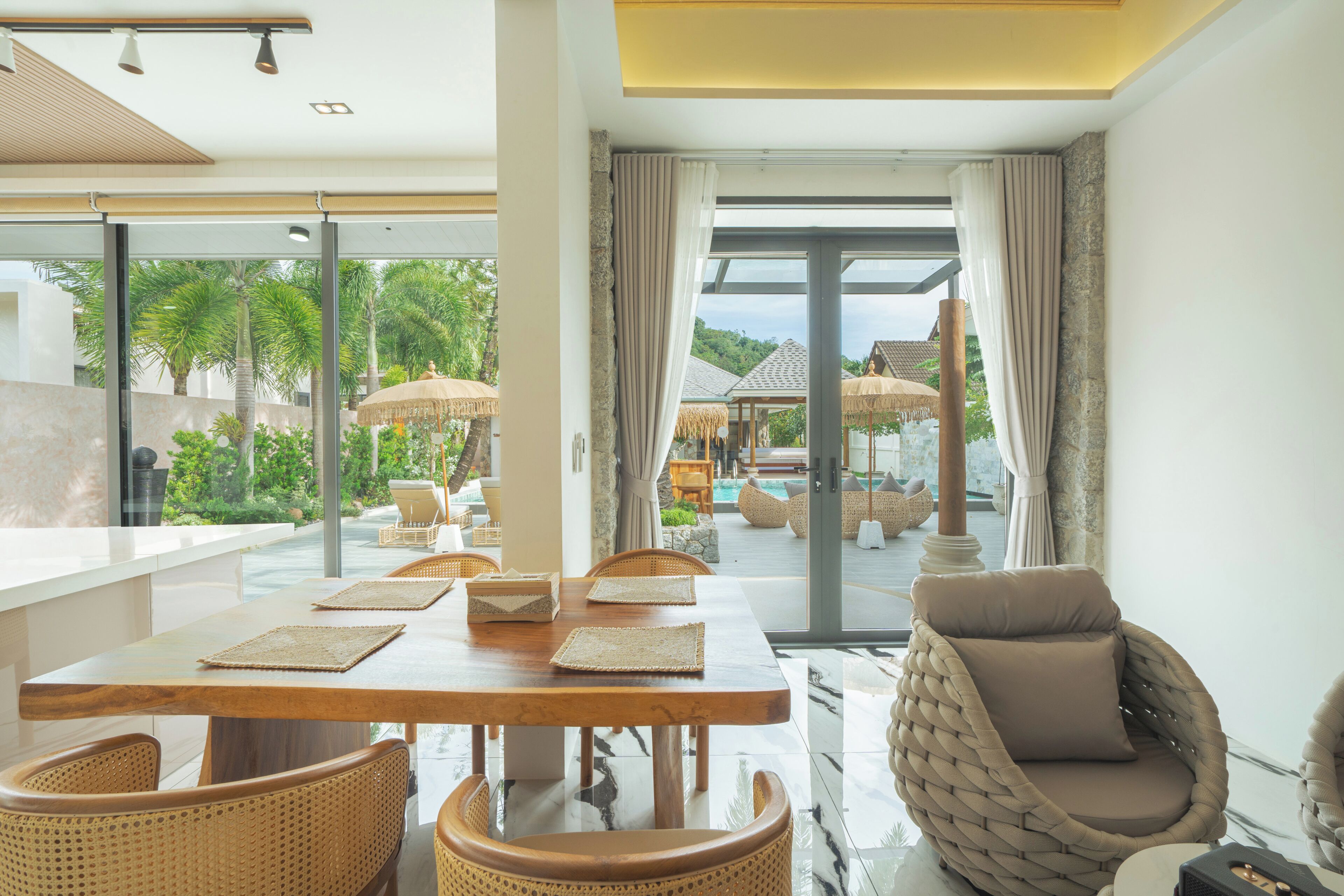 A modern dining area with a wooden table, woven chairs, and a view of a pool and outdoor seating.