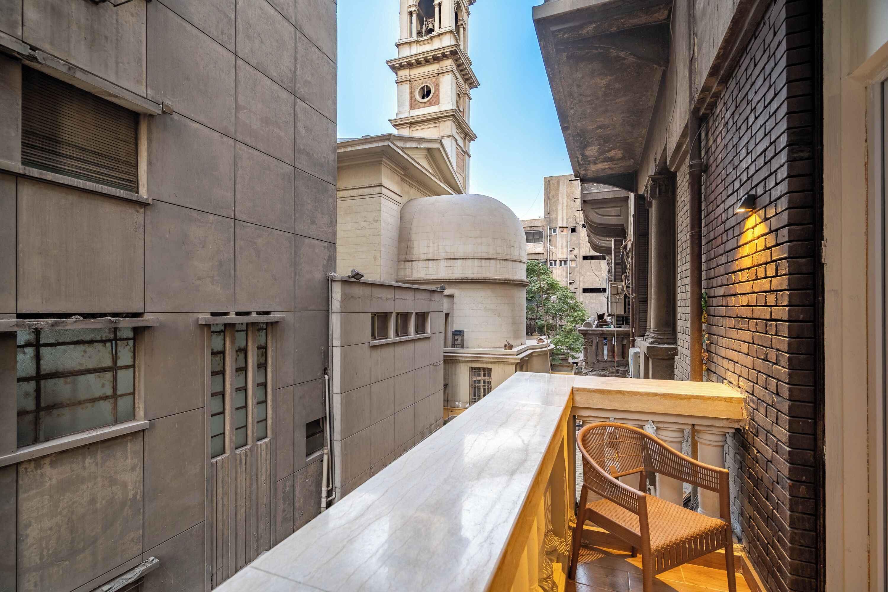 A narrow alleyway with a bar table and chairs, a brick wall, and a building with a dome and bell tower in the background.