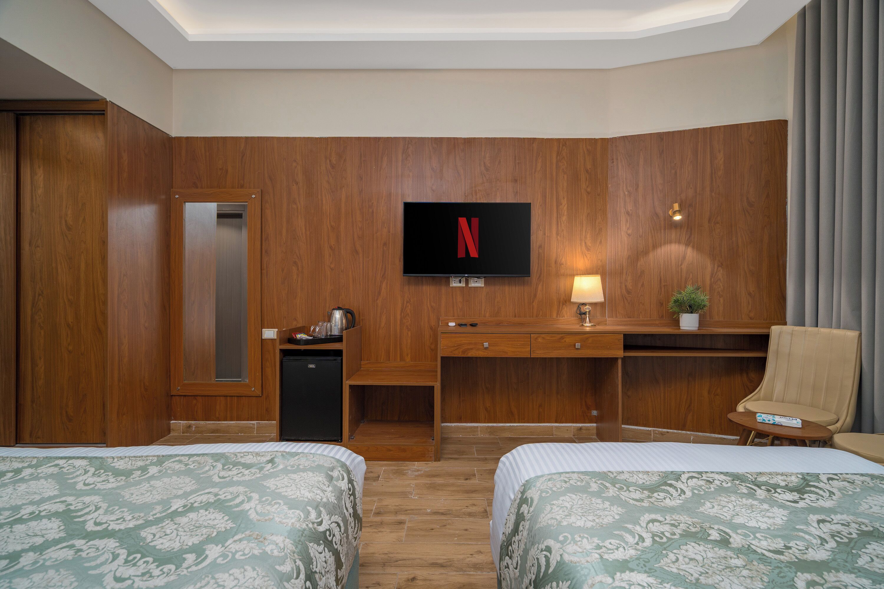 Une chambre d’hôtel avec deux lits, un bureau, une télévision, un petit réfrigérateur, une lampe et une plante en pot.