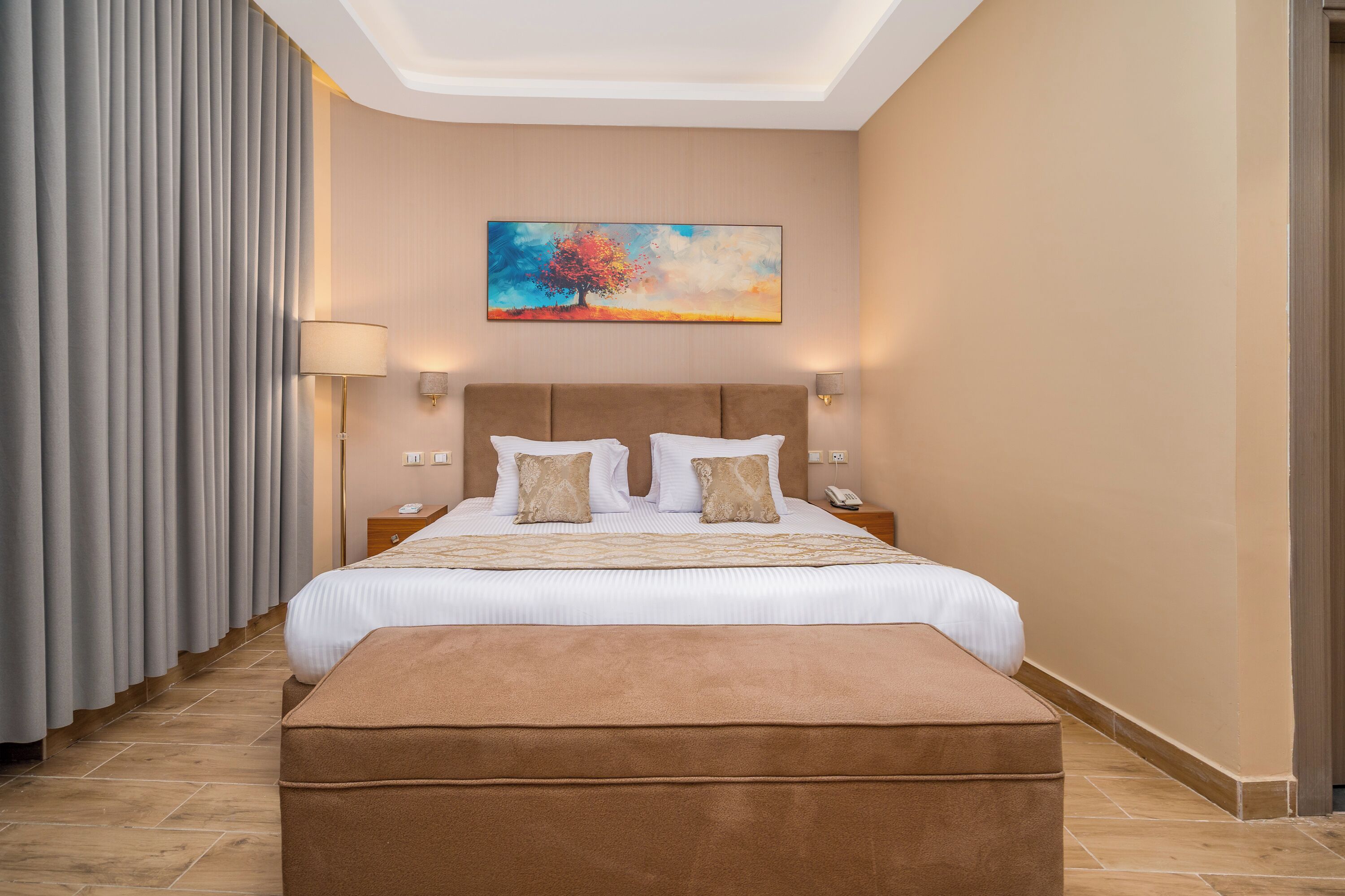 A hotel room with a large bed, a bedside table with a lamp, a painting on the wall, and a window with curtains.