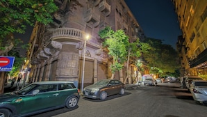 Uma rua com carros estacionados, um edifício com arquitetura elaborada e uma placa de proibido estacionar.