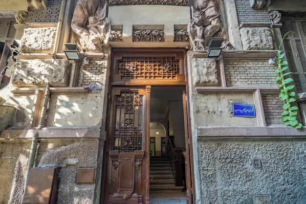 A building entrance with a wooden door, a staircase leading inside, and a blue sign on the wall.