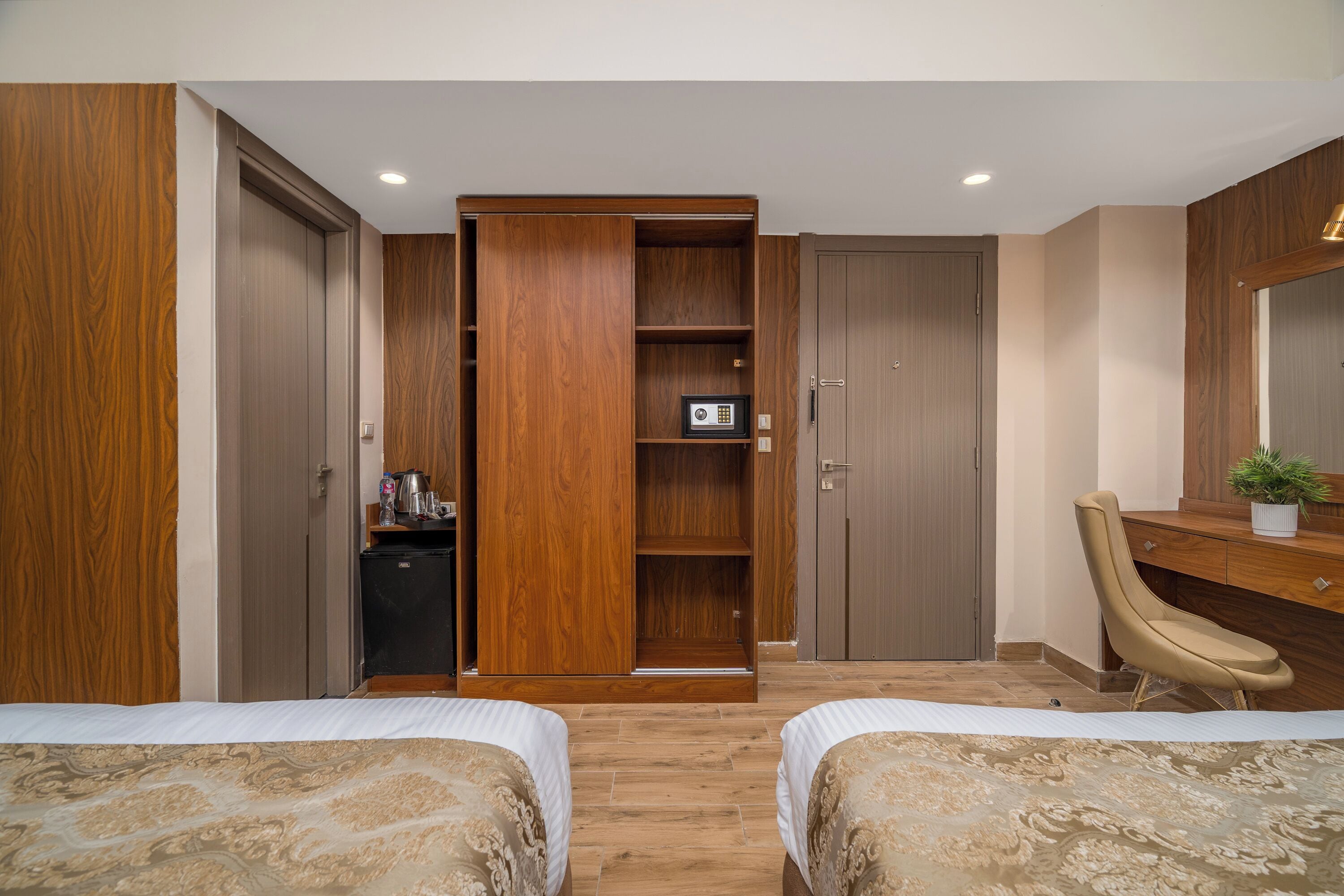 A hotel room with two beds, a wardrobe, a desk with a chair, and a small plant.