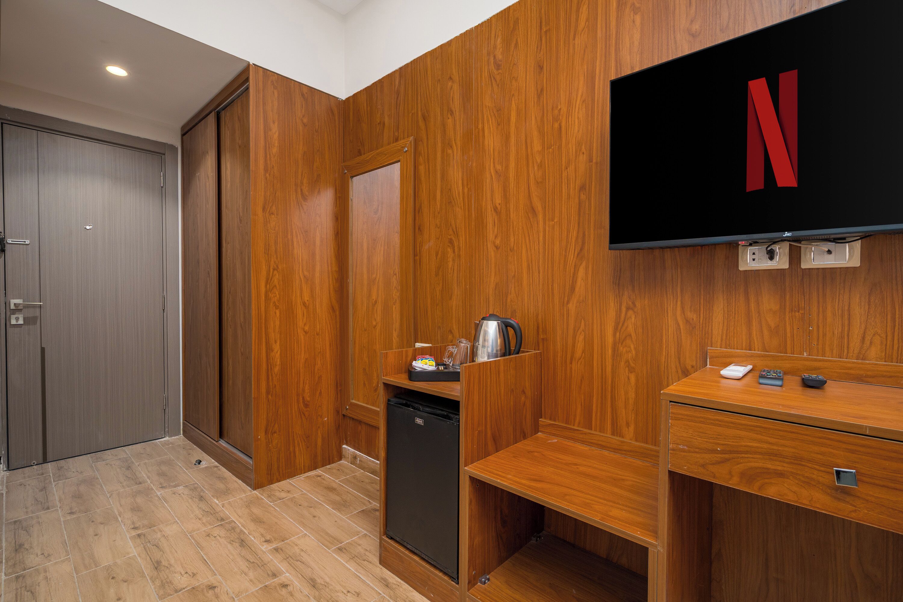 A hotel room with a wooden wardrobe, a small kitchenette, a flat-screen TV mounted on the wall, and a wooden floor.