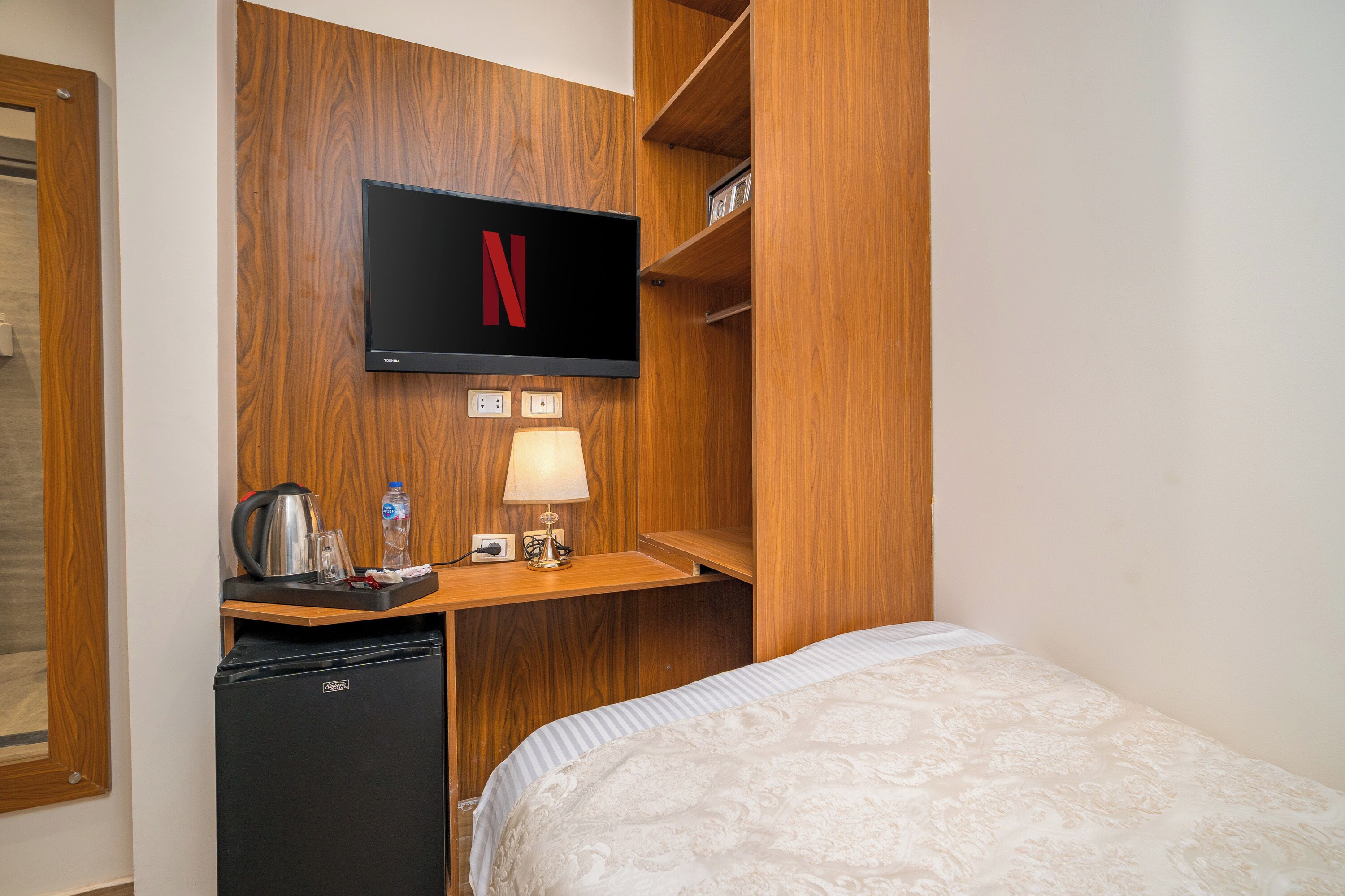 Une chambre d’hôtel avec un lit, un bureau sur lequel se trouve un téléviseur affichant le logo de Netflix, une bouilloire, une bouteille d’eau et une lampe.