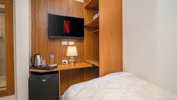 Une chambre d’hôtel avec un lit, un bureau sur lequel se trouve un téléviseur affichant le logo de Netflix, une bouilloire, une bouteille d’eau et une lampe.