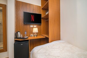 A hotel room with a bed, a desk with a television displaying the Netflix logo, a kettle, a water bottle, and a lamp.