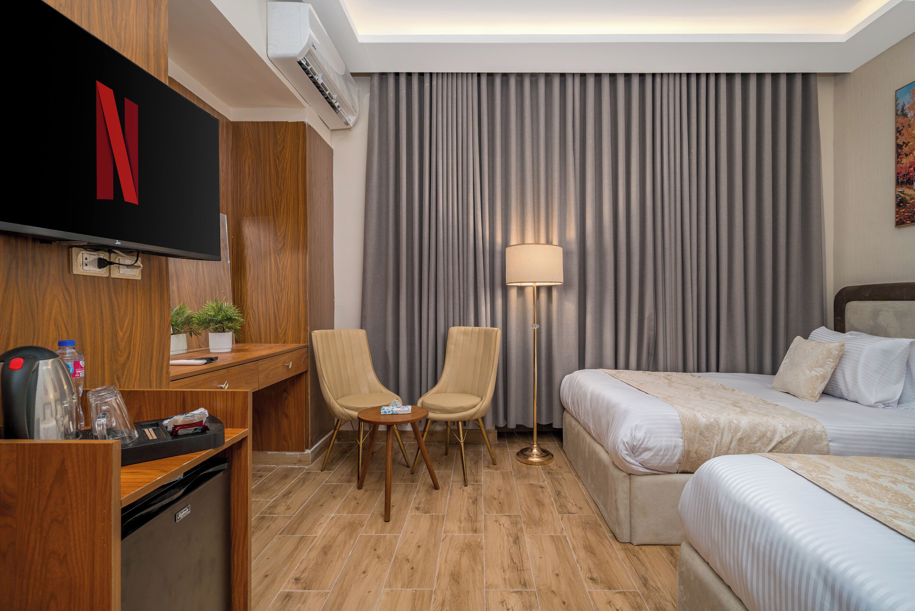 A hotel room with a flat-screen TV, a desk with a chair, a bed, a floor lamp, and a large window with curtains.