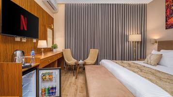 A hotel room with a bed, a desk with a computer, a television, a refrigerator, and a window with curtains.