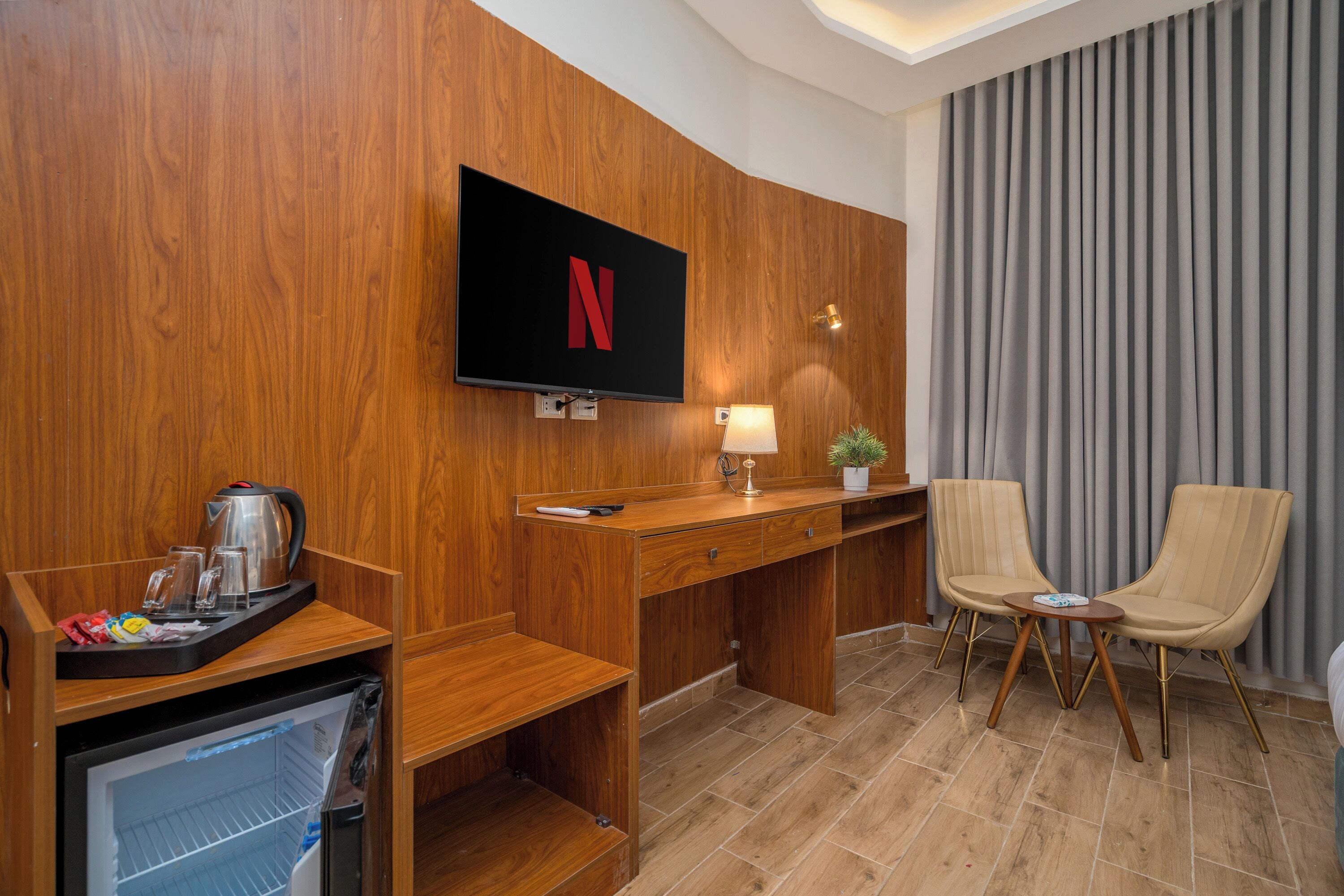 Une chambre d’hôtel avec un mur en bois, un téléviseur à écran plat fixé au-dessus d’un bureau, une lampe, deux chaises et une petite plante en pot.