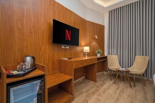 A hotel room with a wooden wall, a flat-screen TV mounted above a desk, a lamp, two chairs, and a small potted plant.