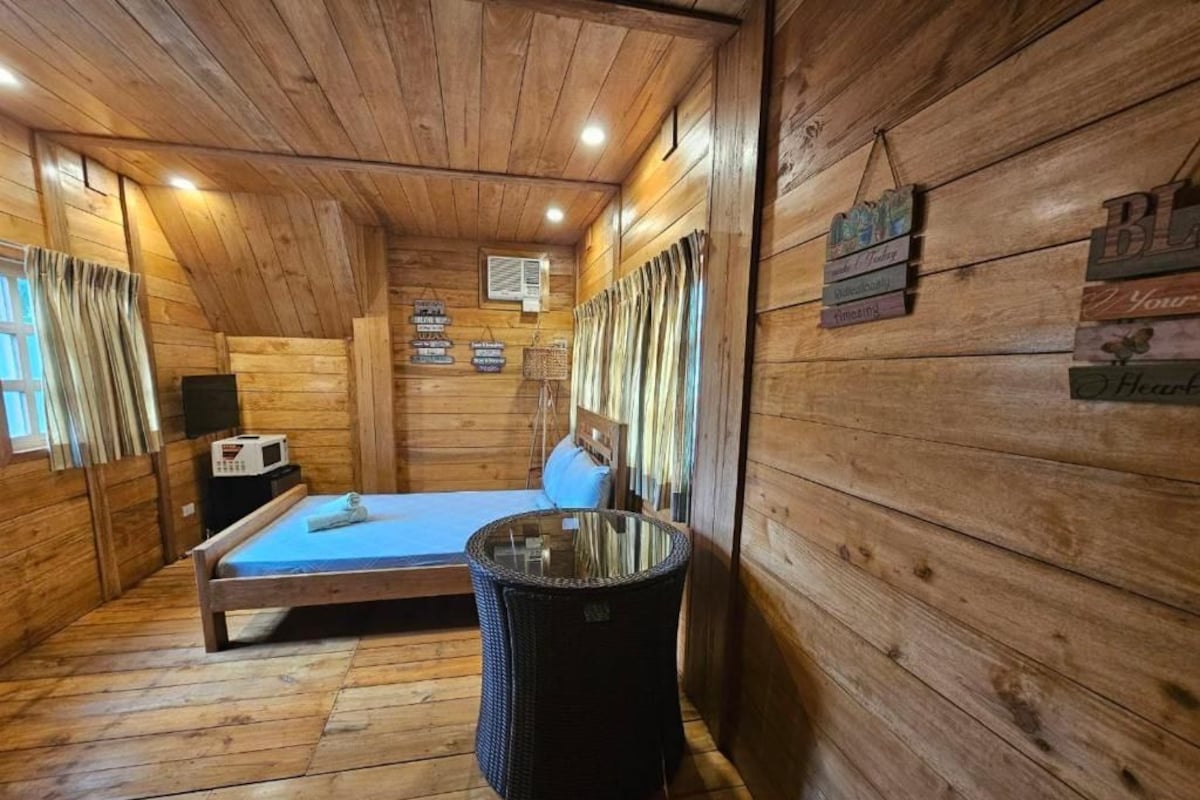 A wooden cabin room with a bed, a small table, and a trash bin.