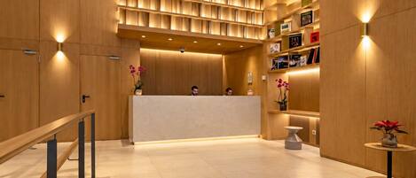A reception area with a wooden reception desk, a seating area with a small table, and a wall with shelves containing books and decorative items.