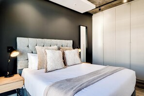 A modern bedroom with a tufted headboard, a bedside lamp, and a large bed with a white comforter. - Mountain View De Waterkant 1 Bed Urban Vue (Cape Town)