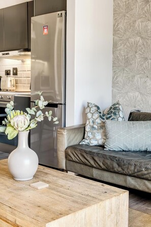 Eine moderne Küche mit Edelstahlgeräten, eine weiße Vase mit Blumen auf einem Holztisch und ein Sofa mit gemusterten Kissen.