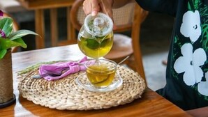 Una persona sirve una bebida en un vaso sobre un posavasos tejido en una mesa de madera, en una habitación con ventanas grandes y vistas a la vegetación exterior.