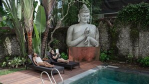 Una estatua de una figura sentada en un jardín con piscina y sillones de descanso.