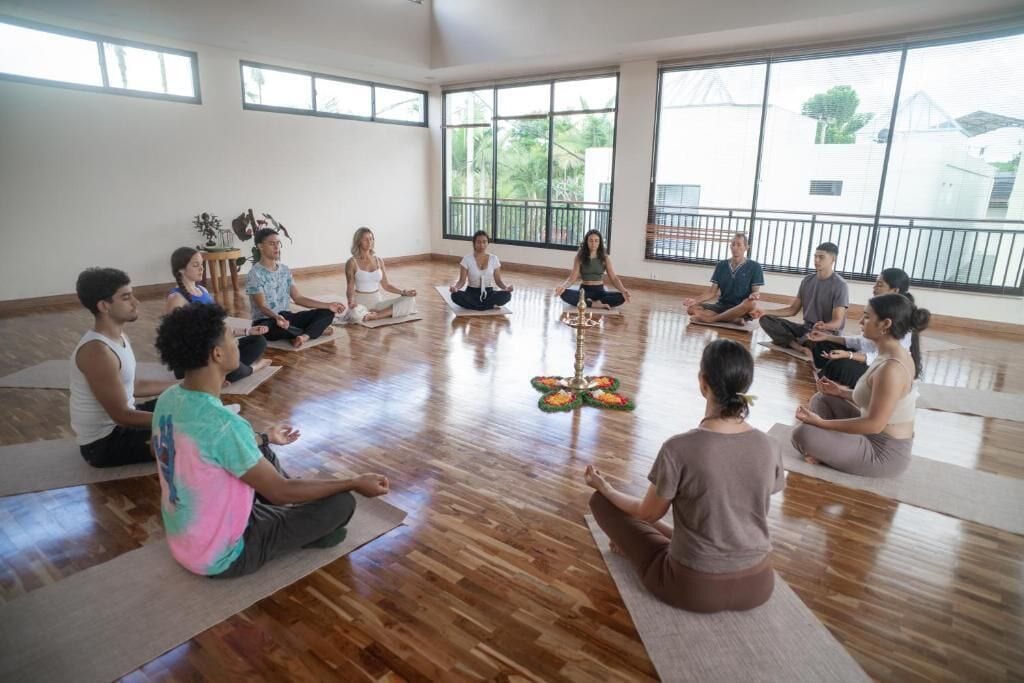 A group of people sitting on yoga mats in a room with large windows and a wooden floor.